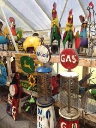 Assorted colorful yard art including roosters and gas pumps displayed on wooden shelves in a greenhouse.