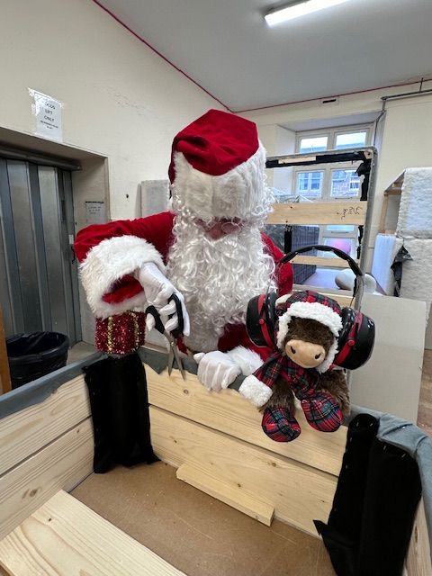 Santa is crafting a bed base from wood, his helper (highland cow) is to his right wearing ear defenders
