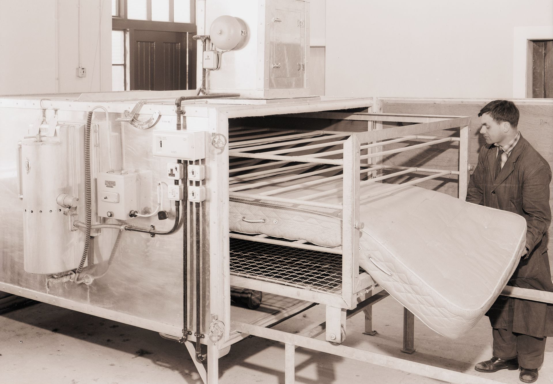 A black and white photo of a man holding a mattress in front of a machine.