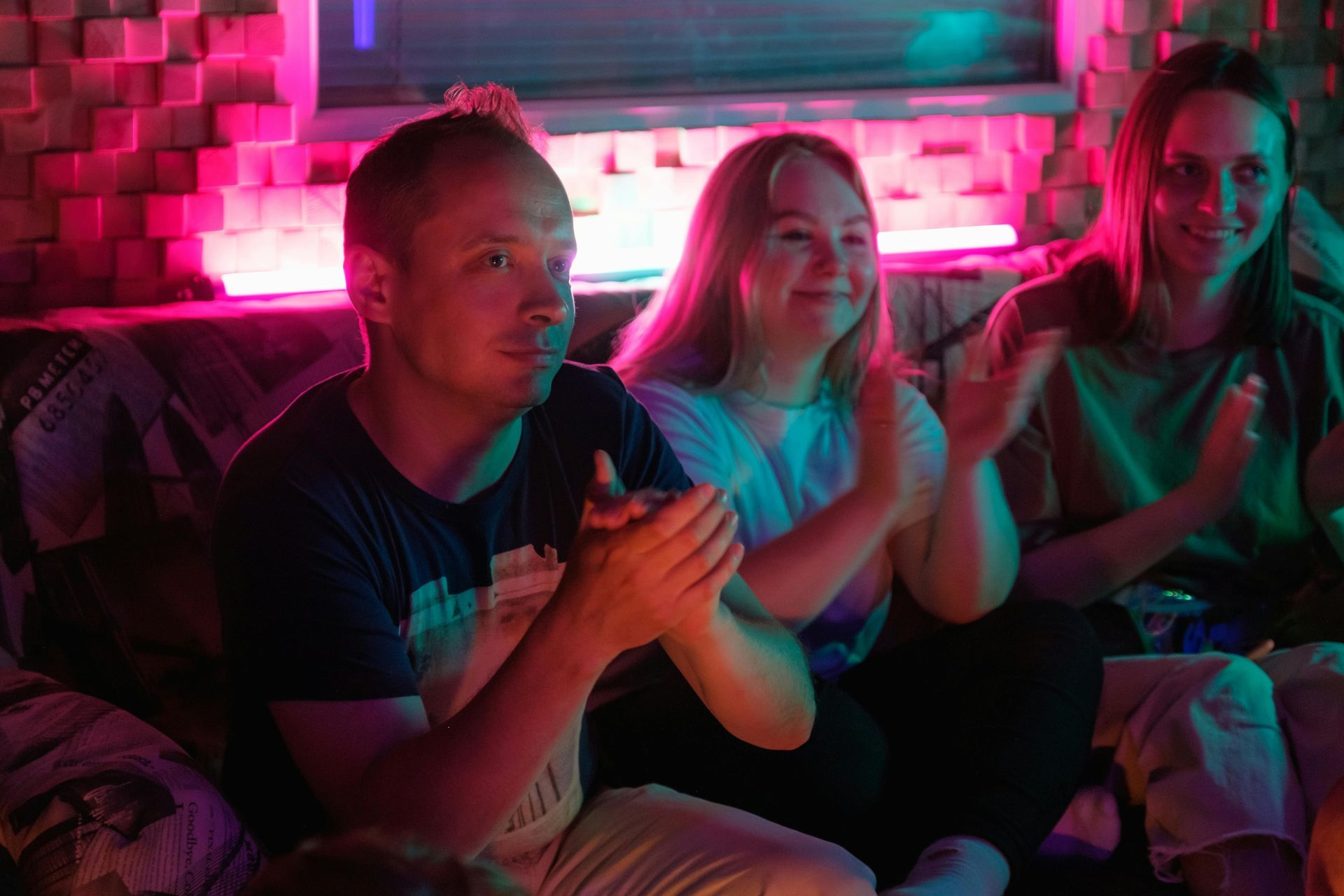 An image of 3 people sitting, there is a man in a black tshirt with blonde hair - a woman in a grey tshirt with blonde hair and a woman in a black tshirt with brown hair.