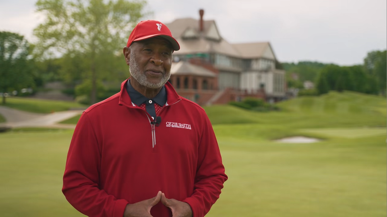 Man in red golf attire on a golf course, hands clasped. Building and green landscape in background.