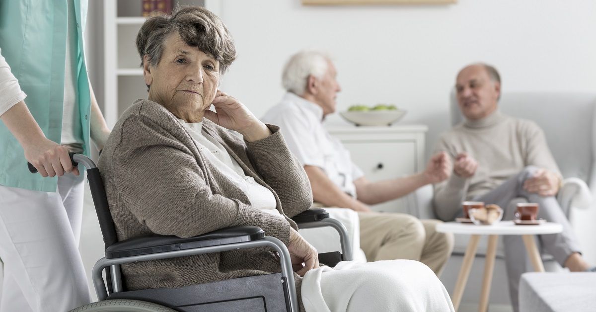 An elderly woman in a wheelchair is sitting in a living room with other elderly people.