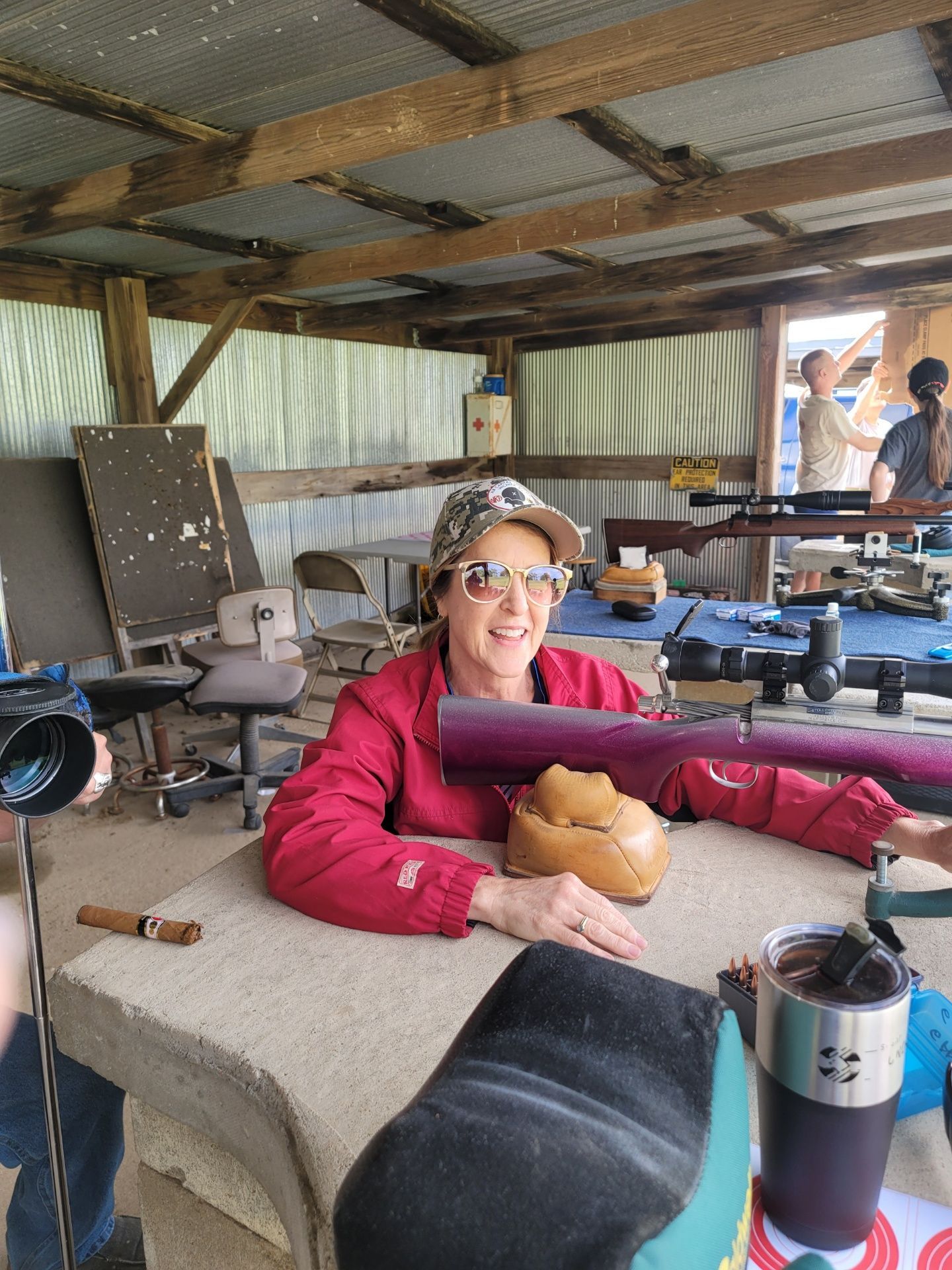 woman in pink jacket wearing a camouflage hat smiling holding a rifle at gun range