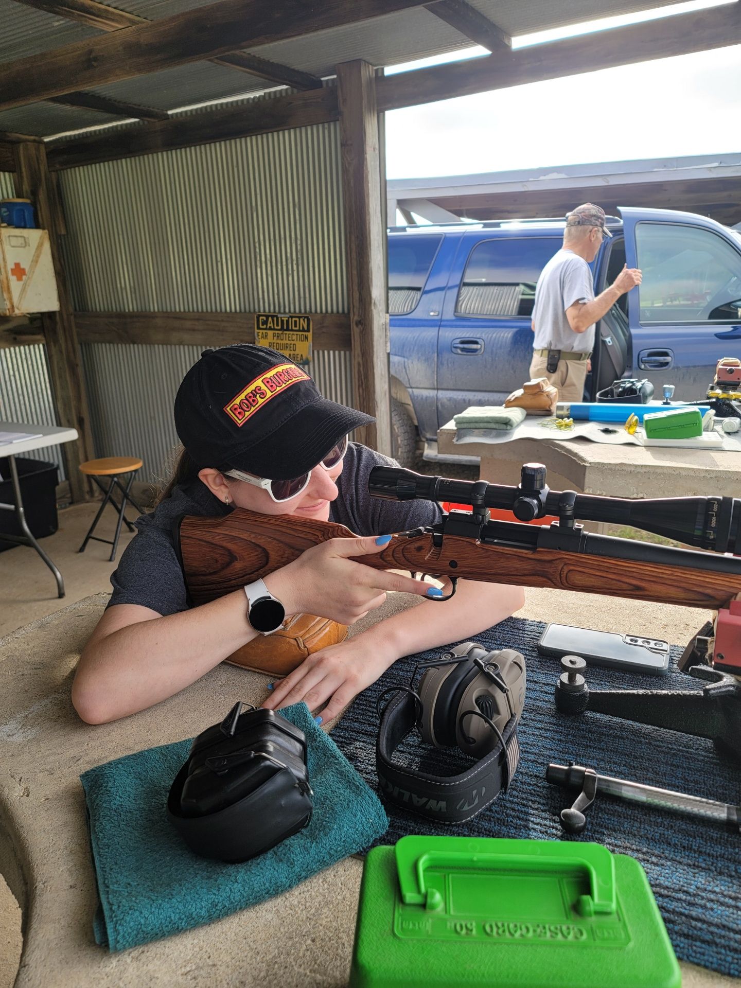 woman in sun glasses and black hat holding a rifle while sitting at gun range table