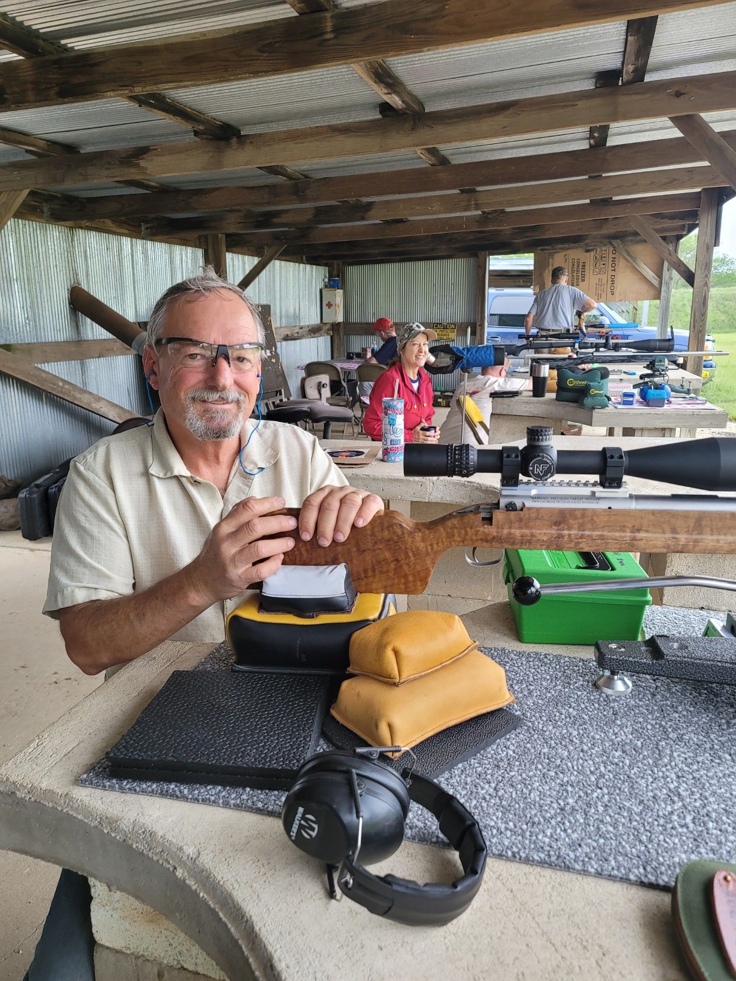 man wearing safety glasses smiling at camera holding a rifle at gun range table