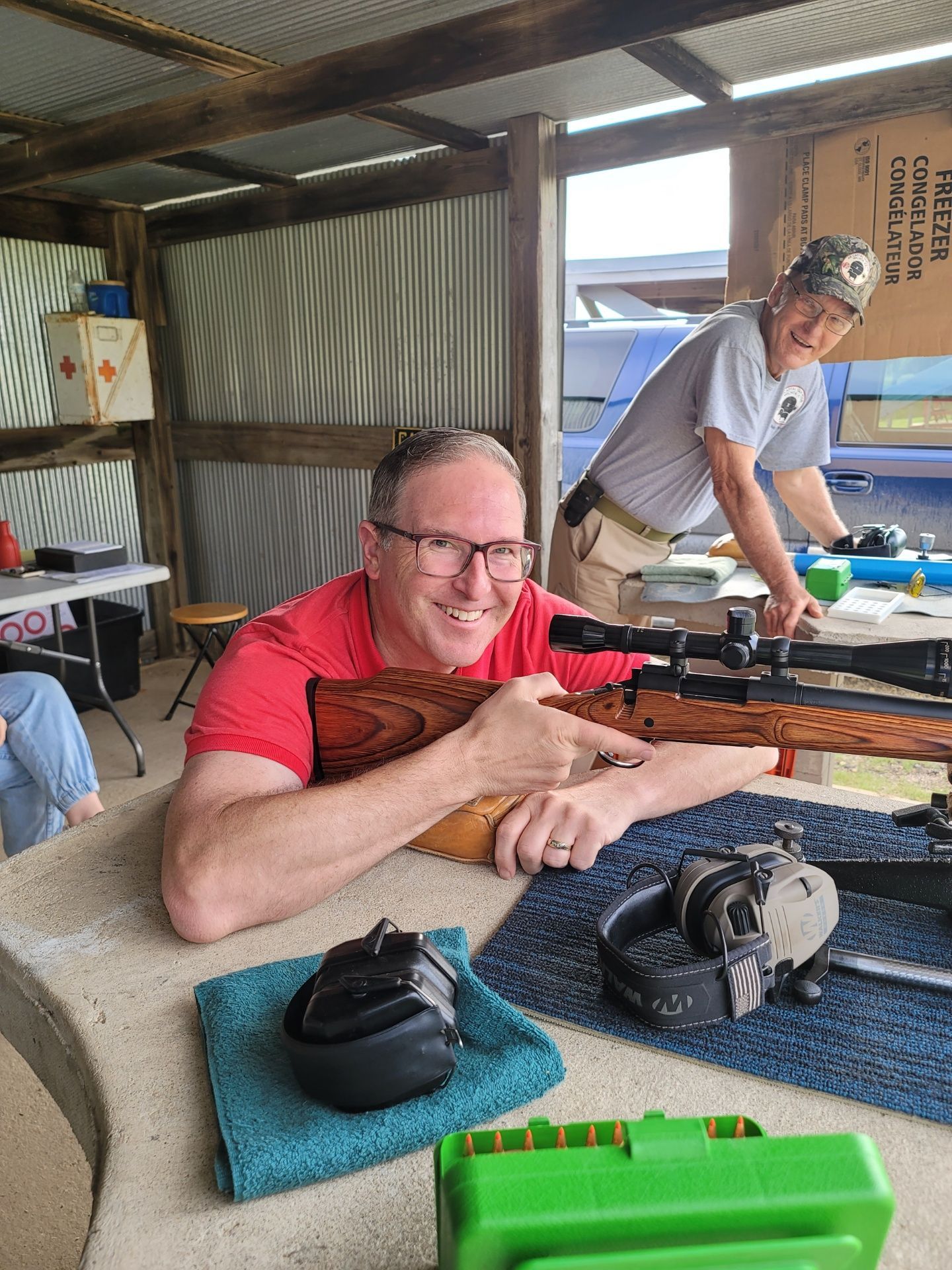 man in red shirt and glasses smiling at camera while holding rifle at a gun range