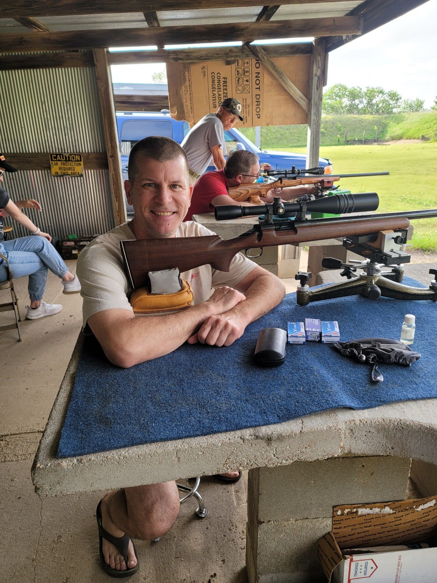 man in tan shirt smiling at camera sitting at gun range table next to rifle