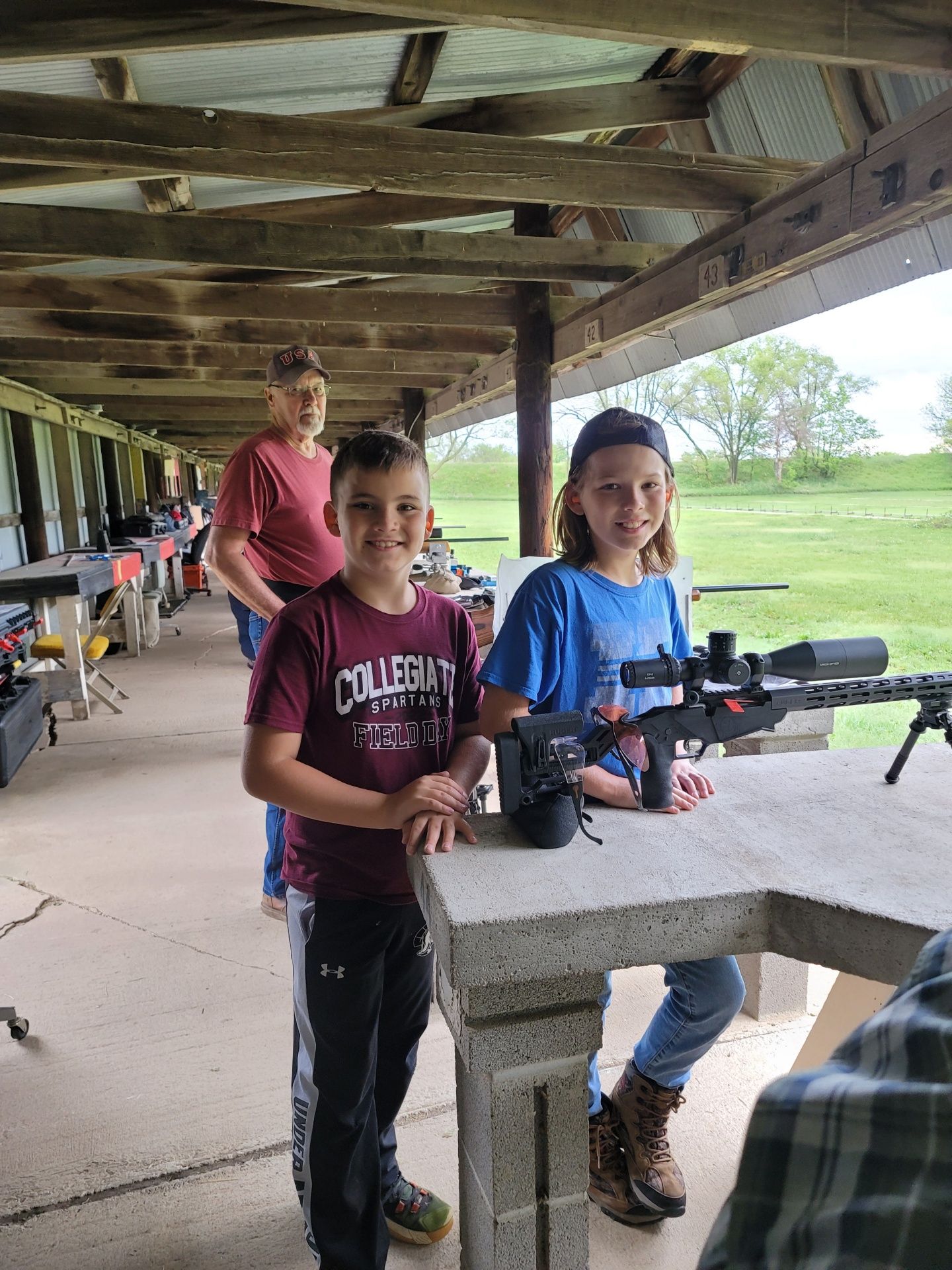 a boy and a girl smiling at the camera while standing by rifle at gun range