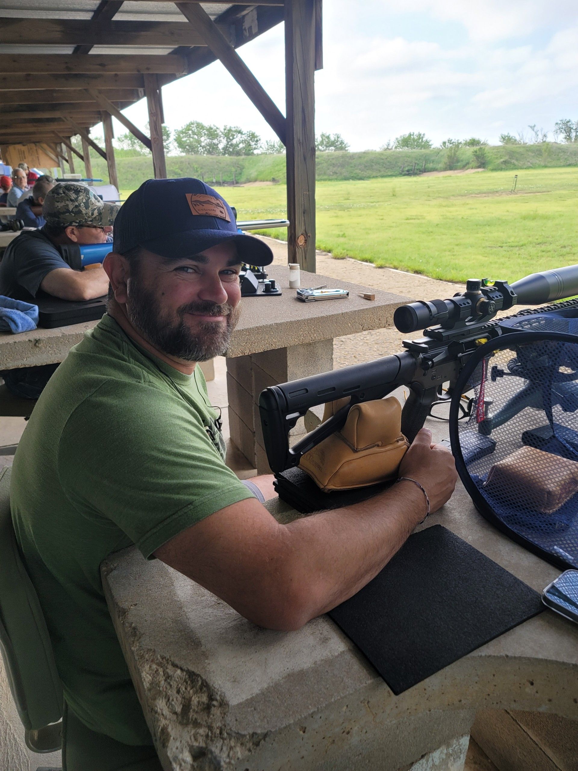man in green shirt and blue hat smiling while sitting at gun range holding a rifle