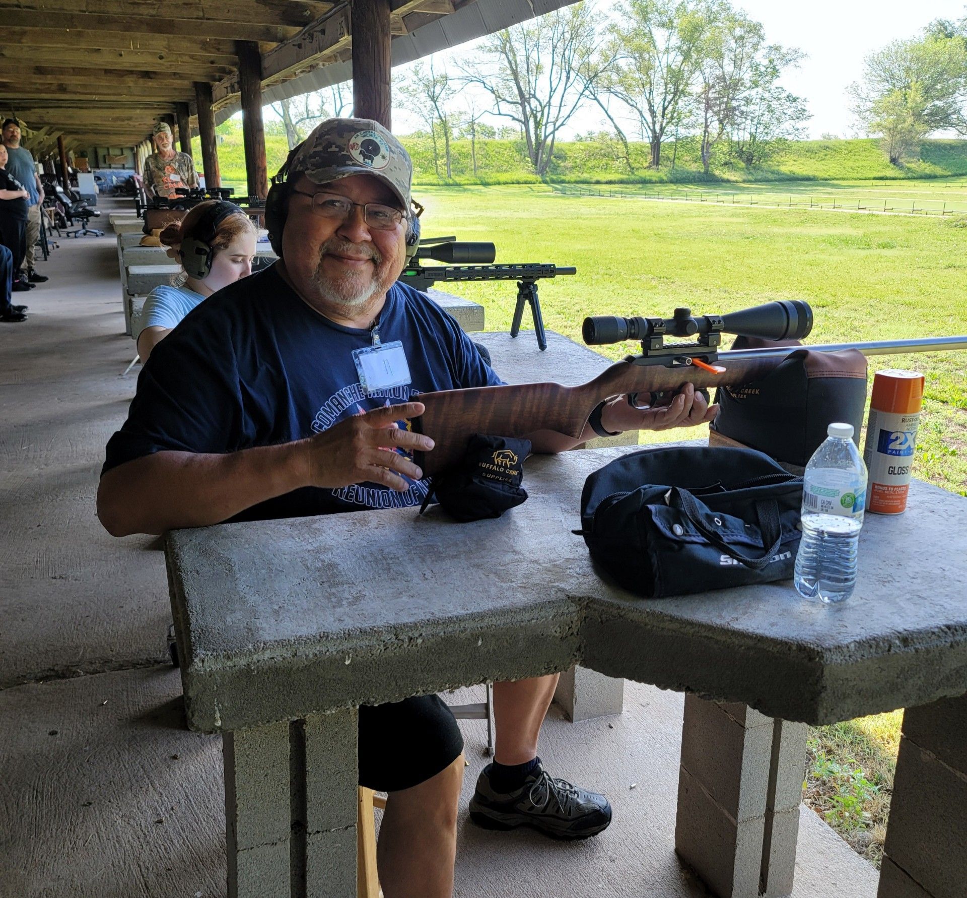 man in blue shirt smiling at camera sitting with rifle at gun range table