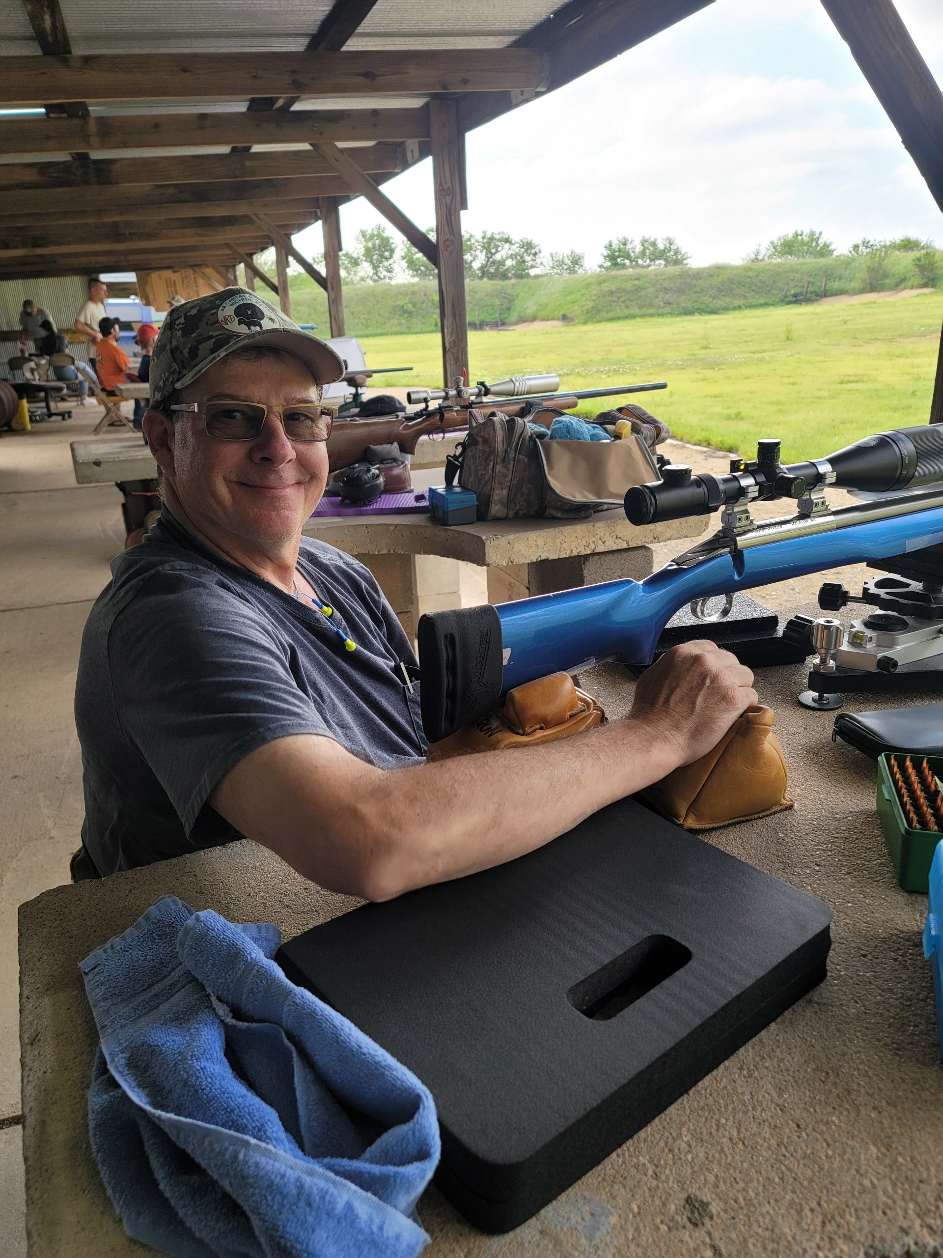 man in blue shirt and hat smiling at camera sitting at gun range table next to rifle