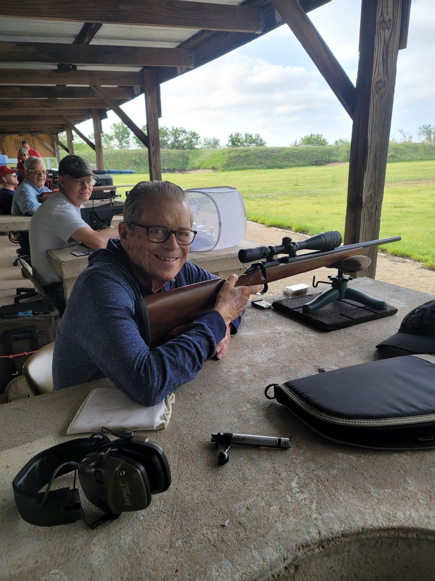 man in blue shirt and glasses smiling at camera sitting at gun range table holding a rifle