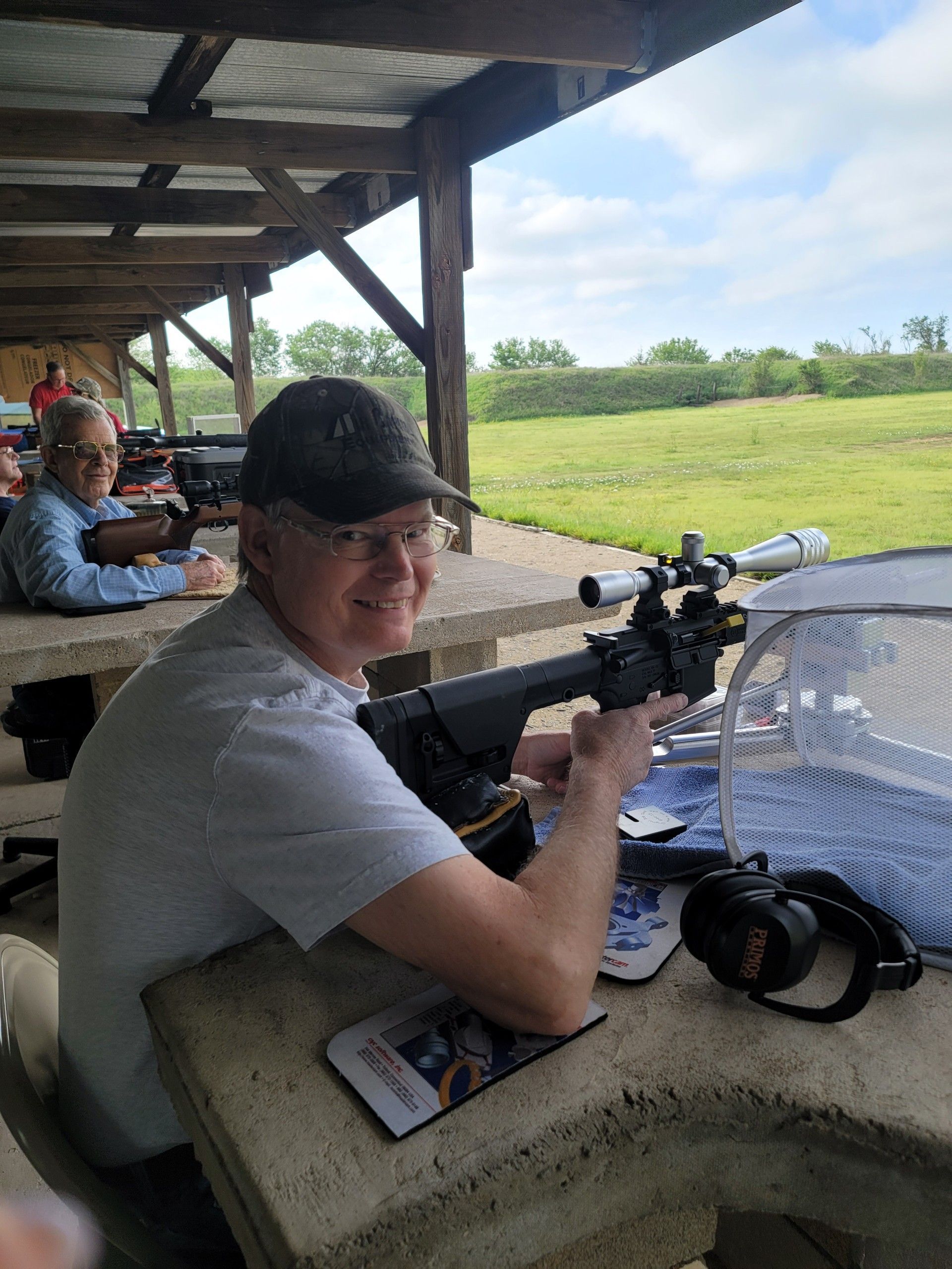 man in glasses and black hat smiling at camera sitting at gun range holding a rifle