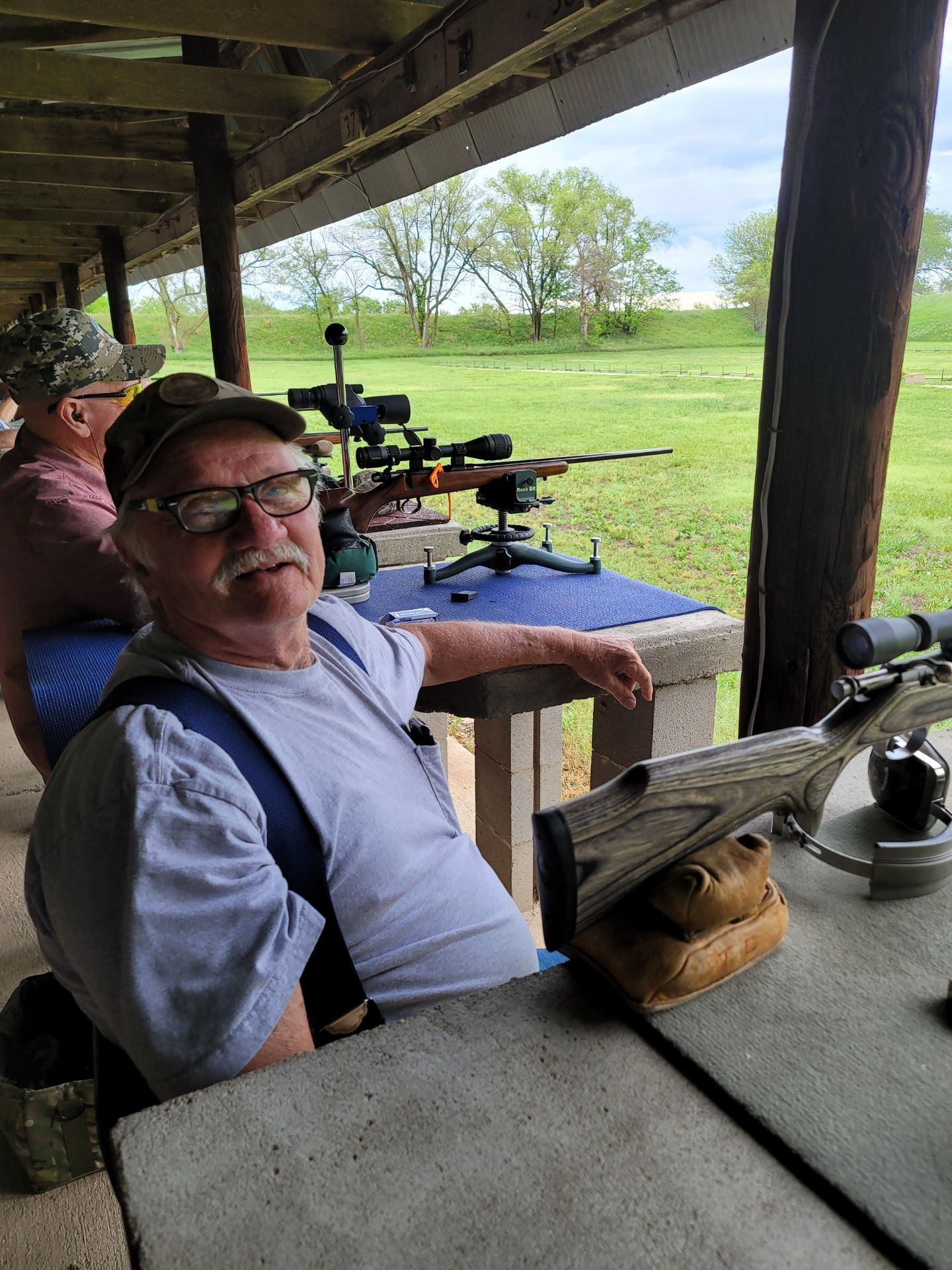 man sitting in gray shirt and glasses at gun range rifle table