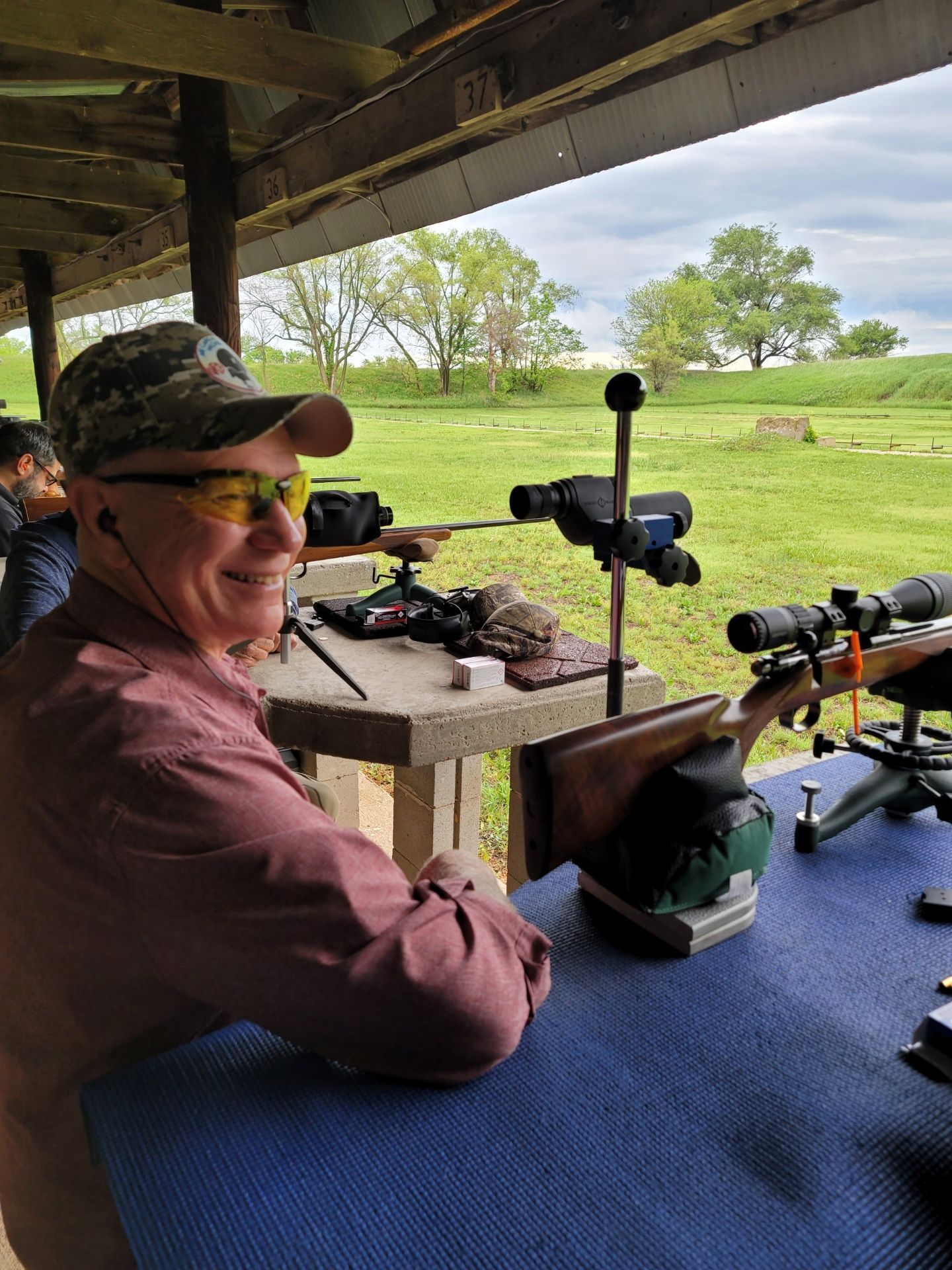 man wearing yellow safety glasses and hat smiling at gun range table