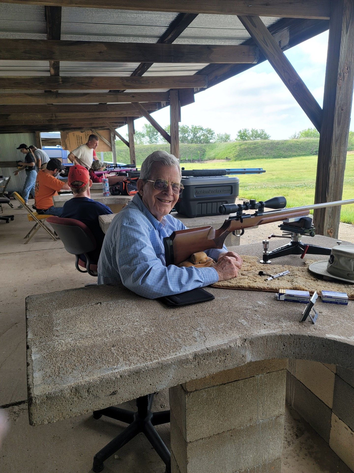 man in blue shirt and glasses smiling at camera sitting at gun range table next to rifle