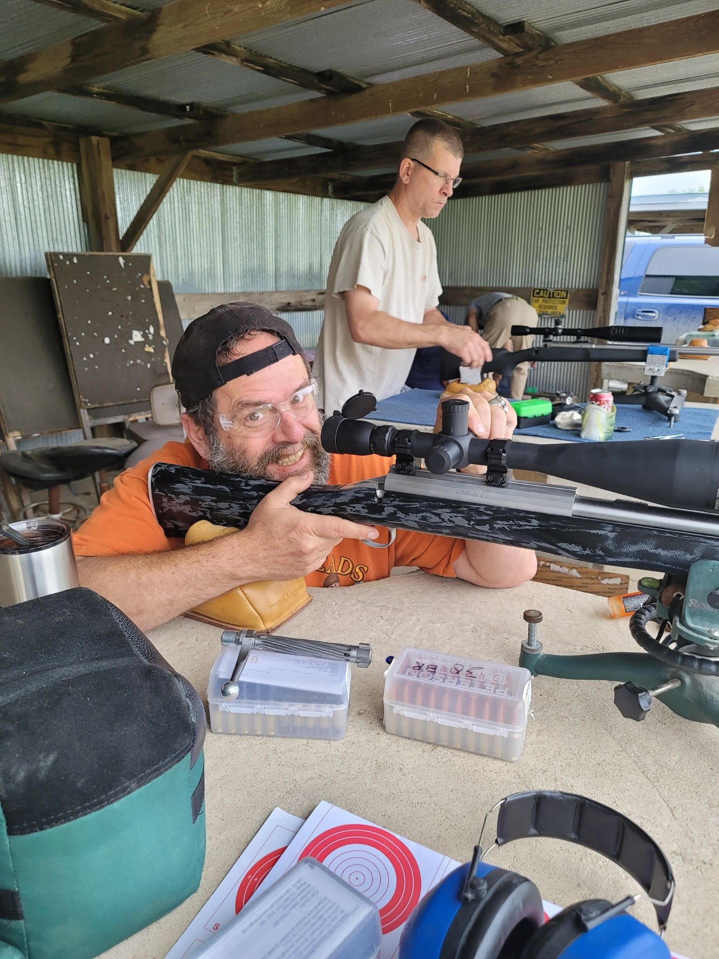 man with safety glasses smiling at camera while holding rifle at gun range table