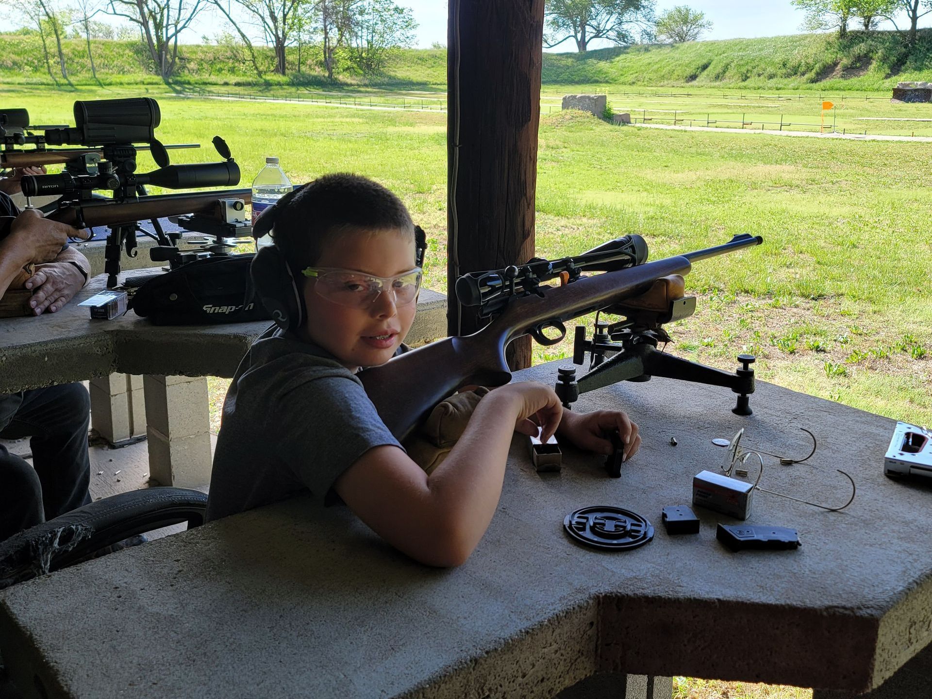 a boy with safety glasses on sitting at gun range holding a rifle