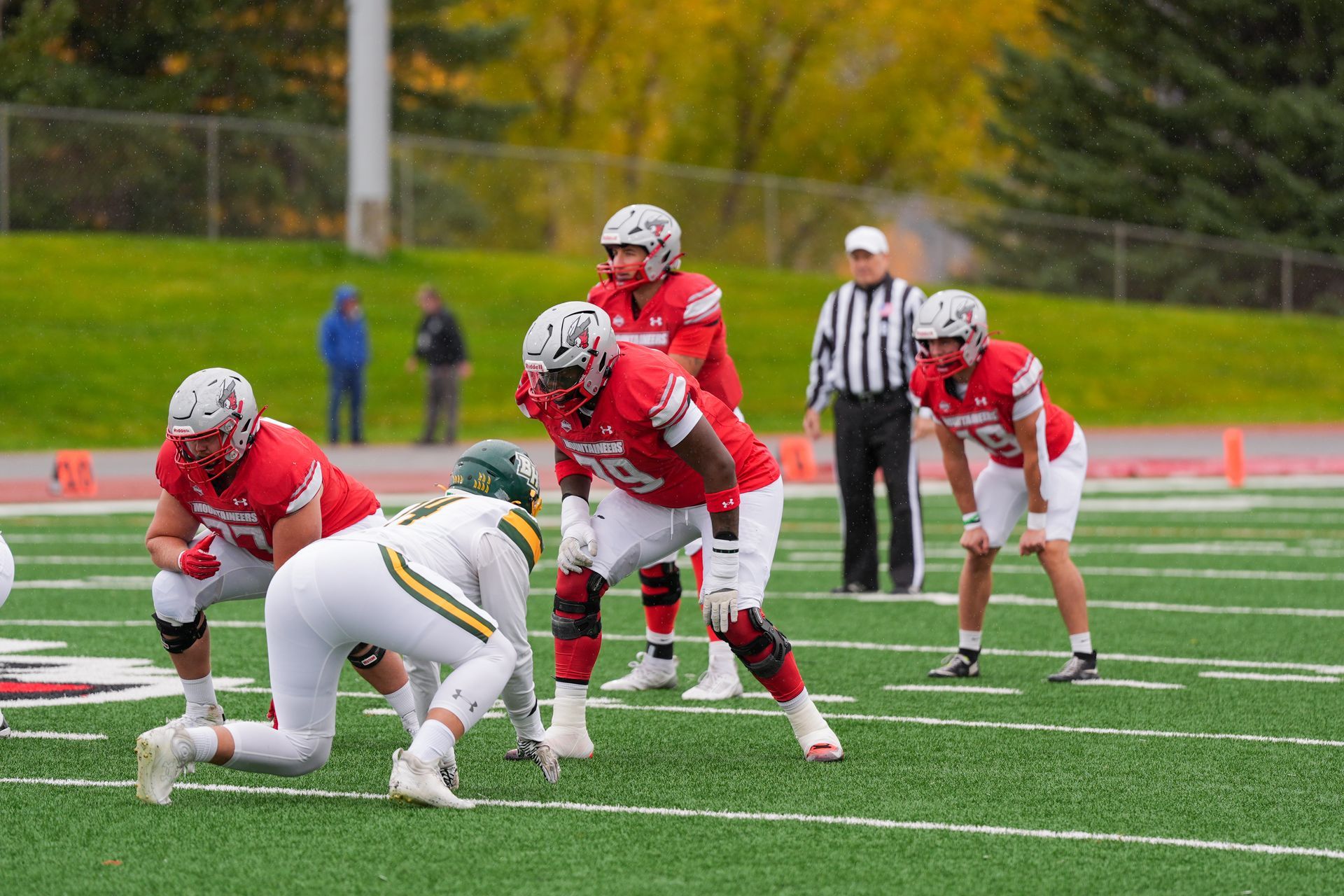 Football players in red uniforms line up against a green-clad player on a field, referee nearby.