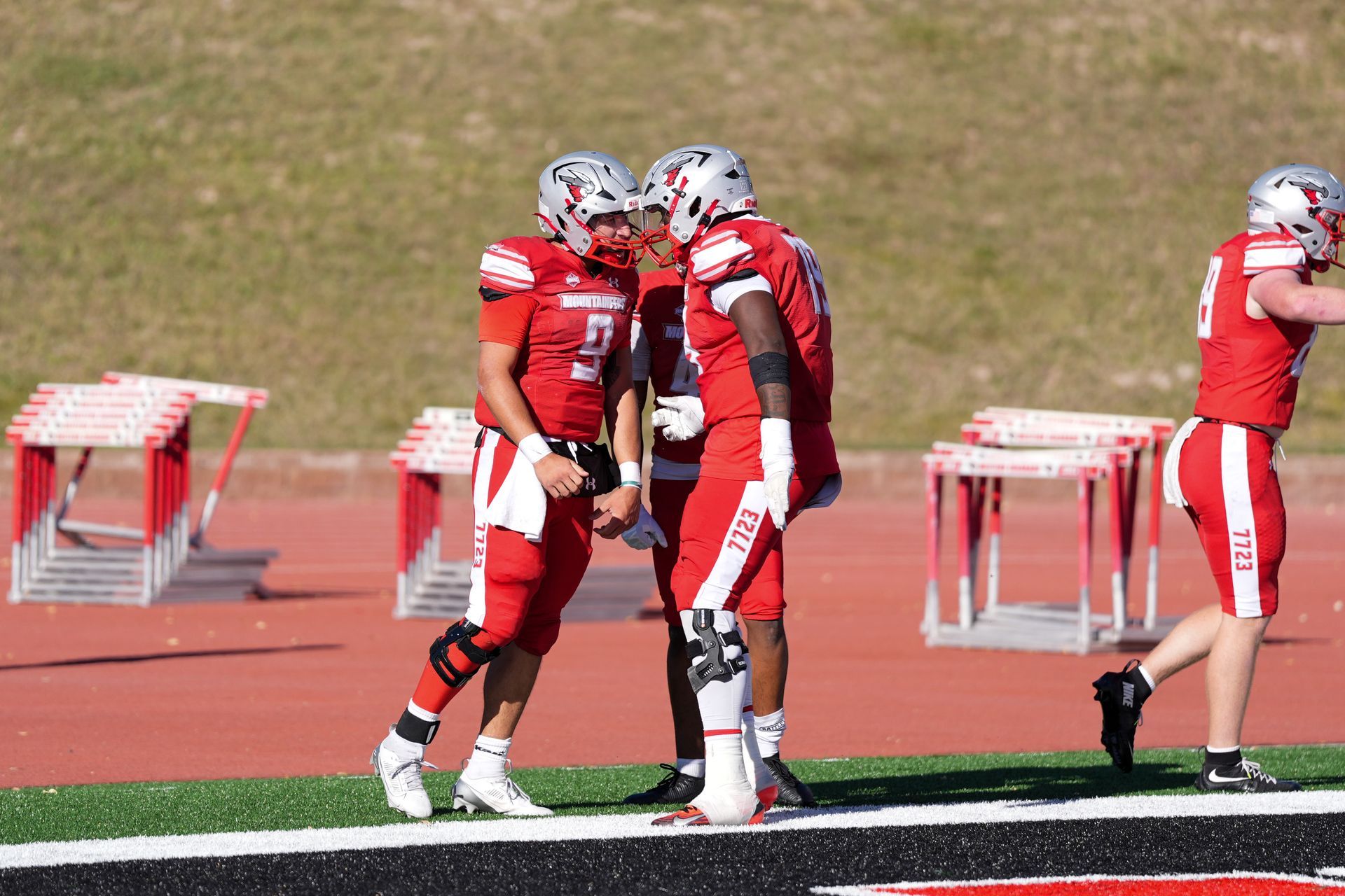 Football players in red uniforms celebrating on a field, near the sidelines with hurdles in the background.
