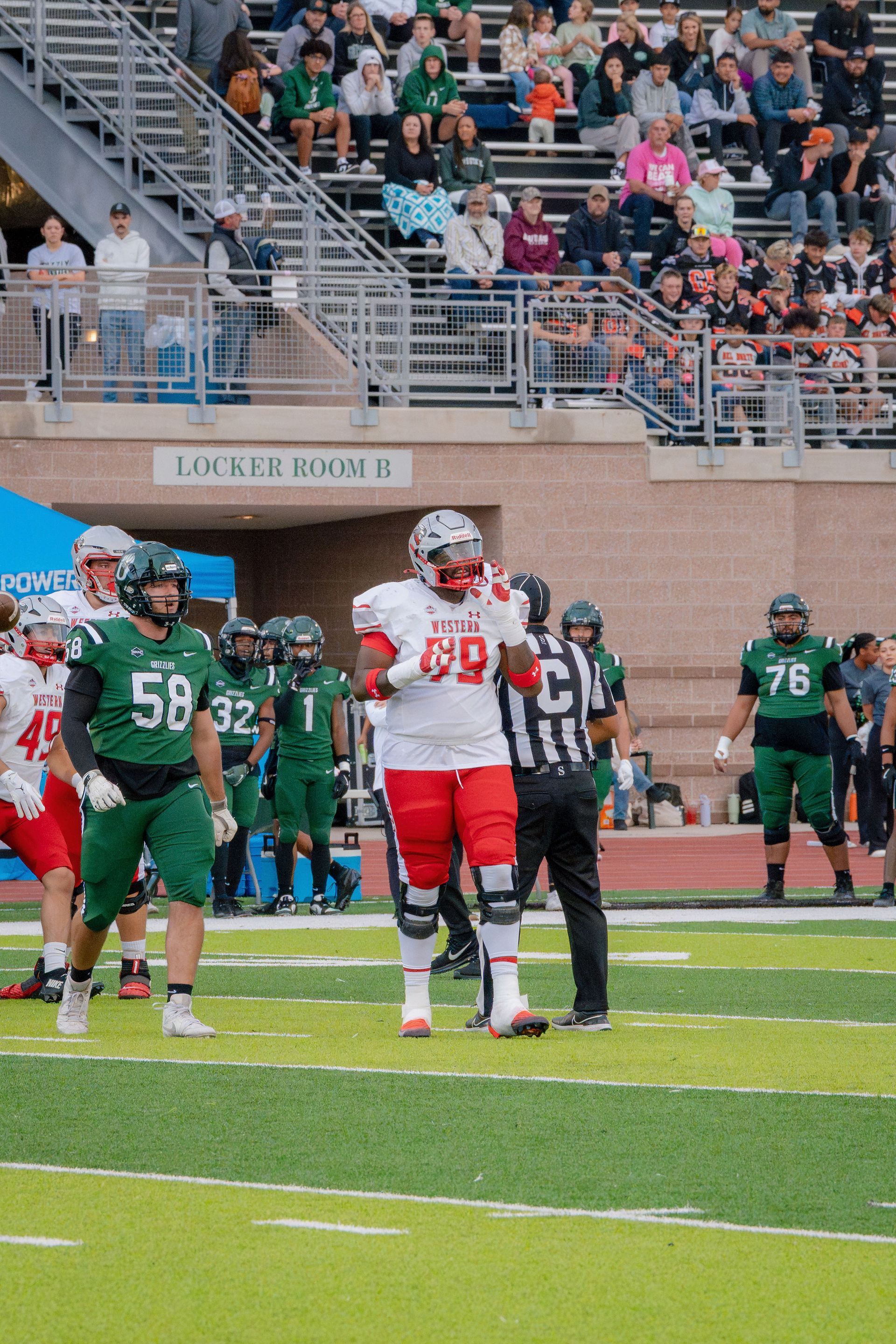Football player in red uniform on a field, others in green, referee nearby, stadium background.