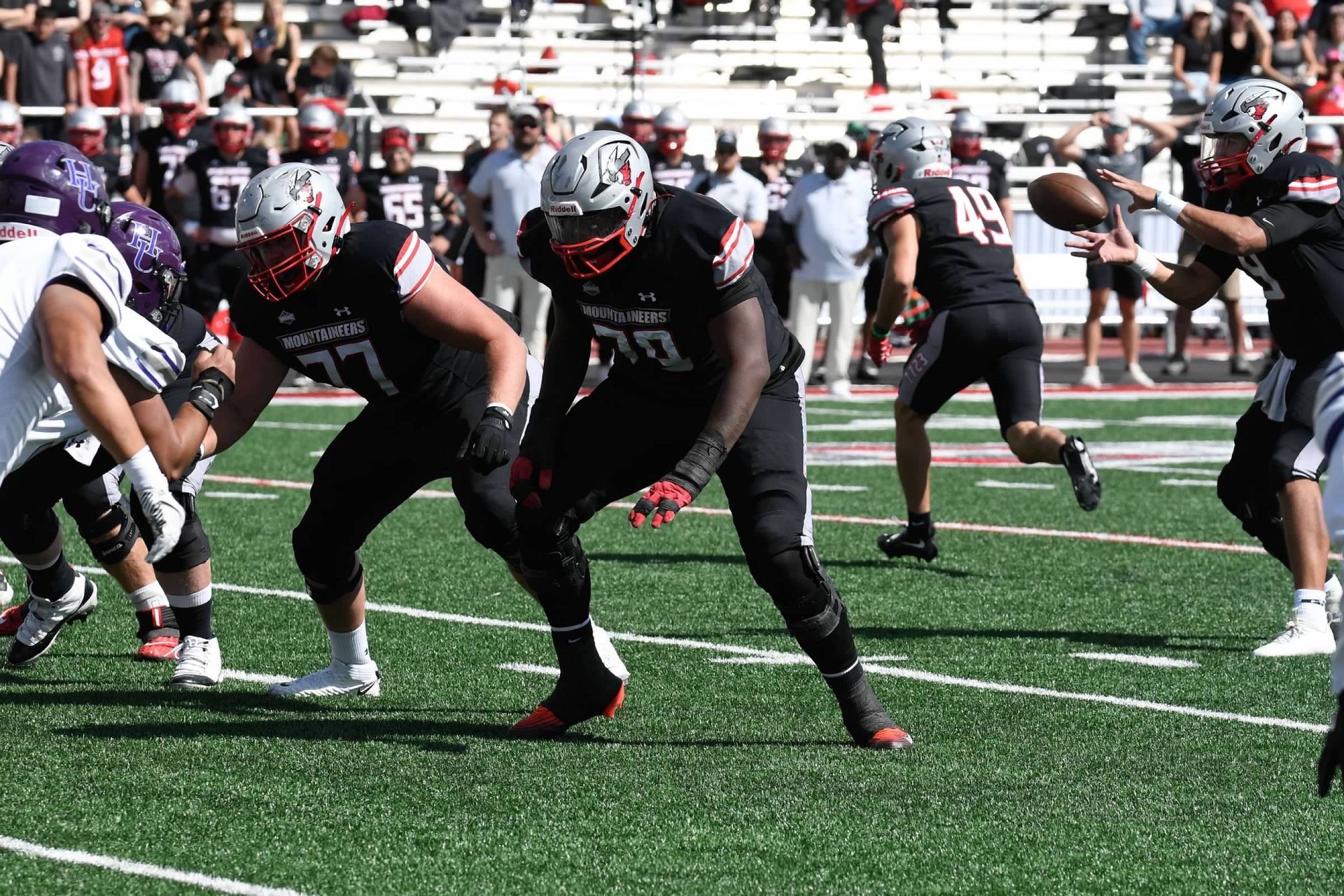 Football players in black uniforms on a green field. One player throws a football, other players in the foreground.