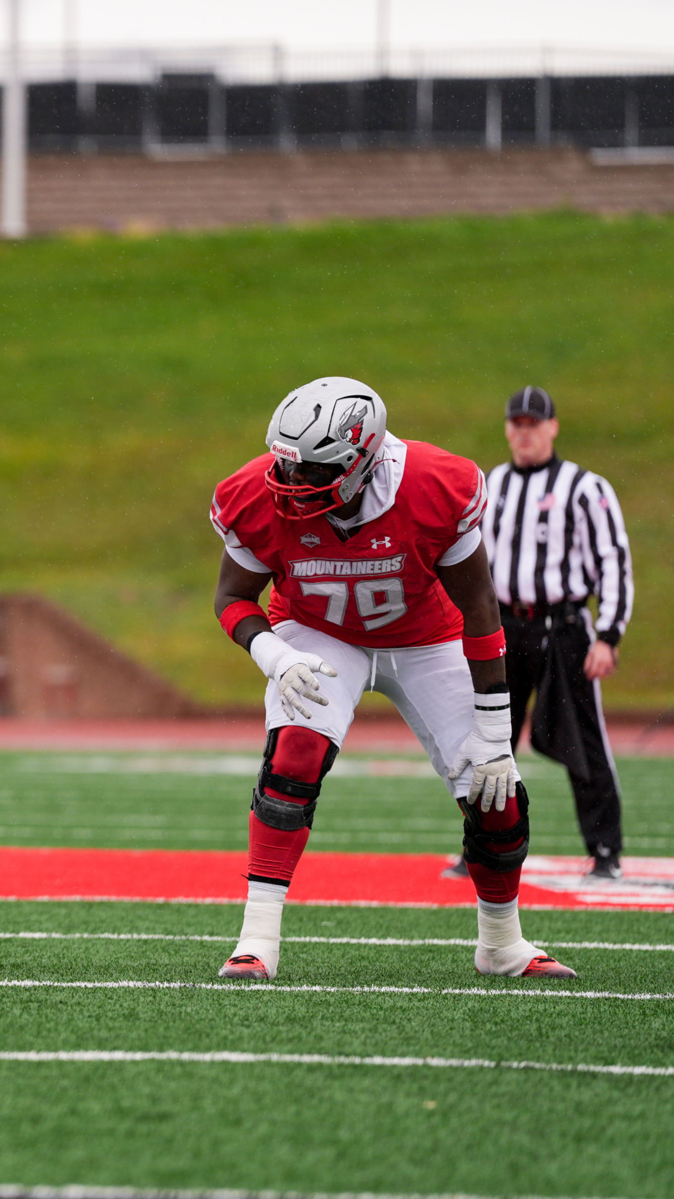 Football player in red uniform, number 79, in stance on a green field. Referee in background.