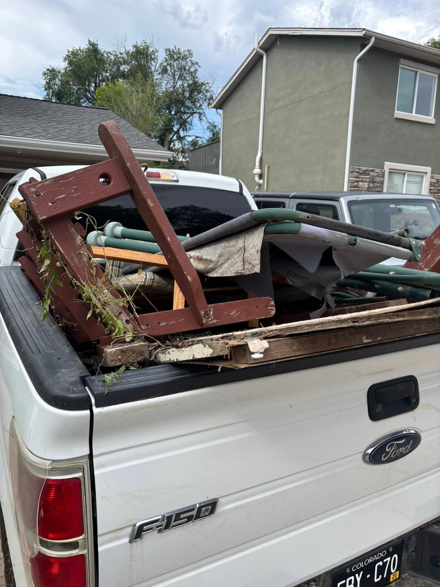 White pickup truck bed filled with debris: wood, fabric, and green tubes. Houses and a cloudy sky in the background.