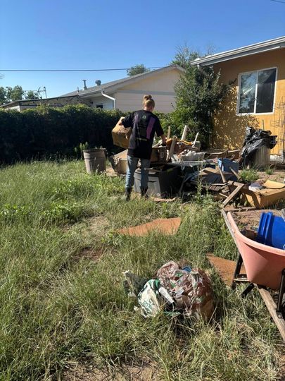 Person in a backyard, carrying a basket towards a pile of objects. Green grass and a yellow house are visible.