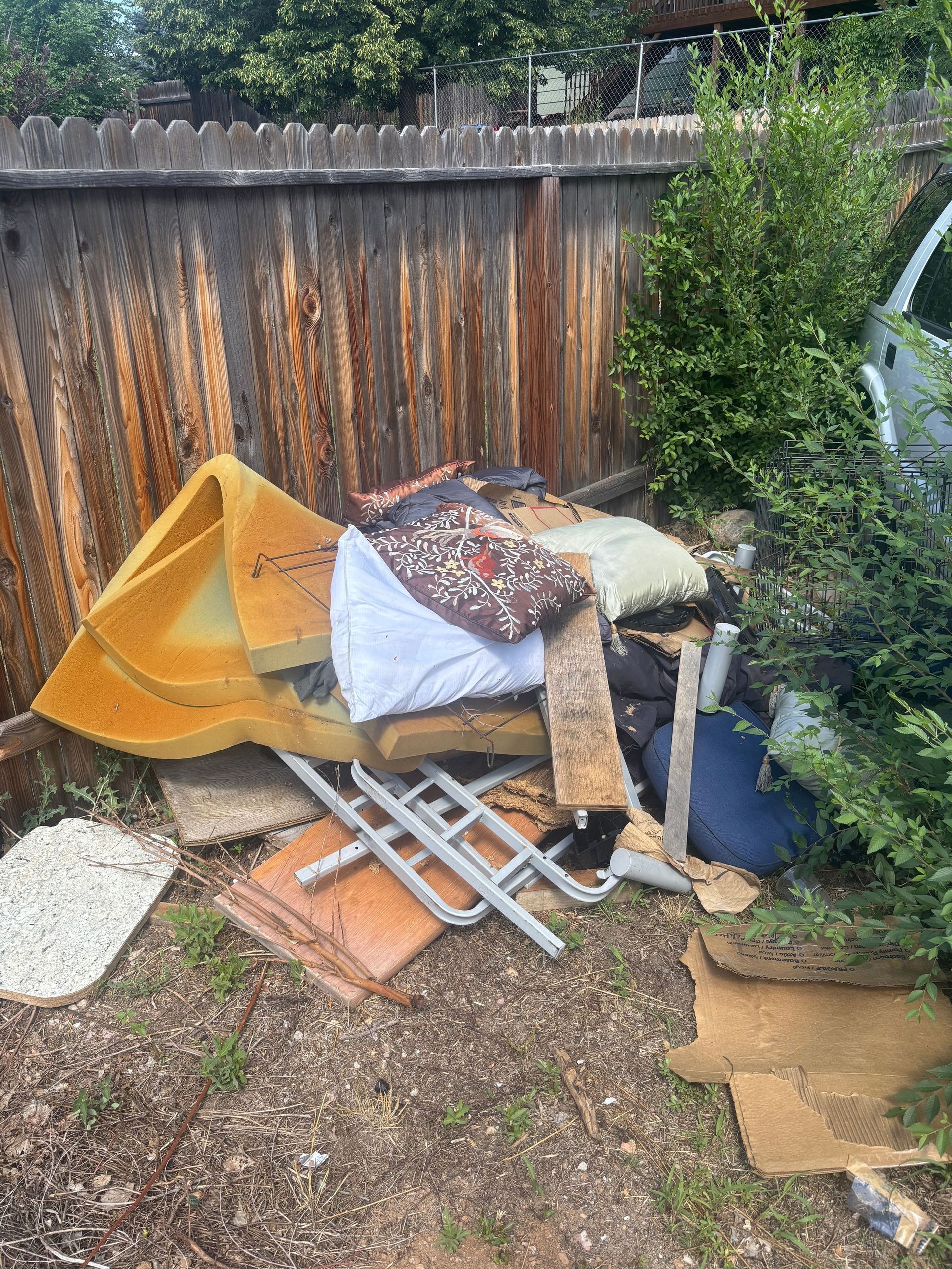 Pile of trash including carpet, cushions, and a metal frame, sits against a wooden fence in a yard.