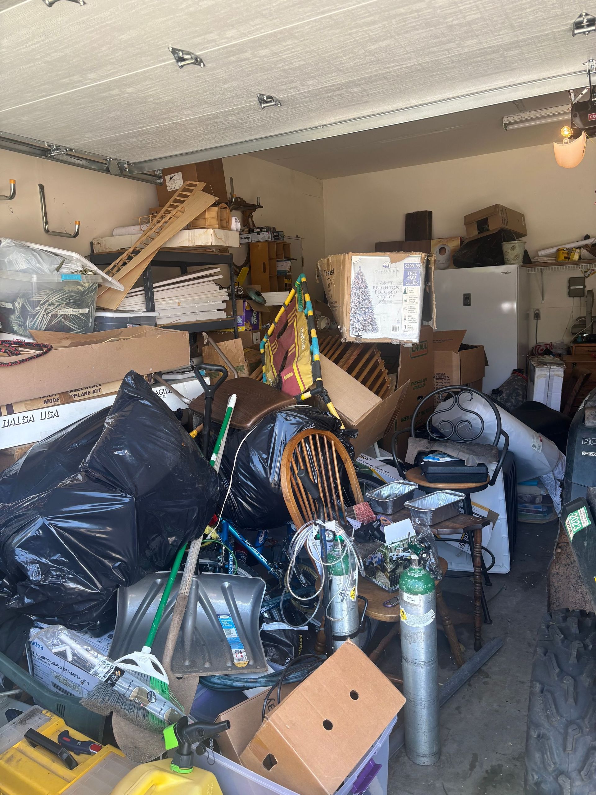 A cluttered garage filled with boxes, bags, and various items, including oxygen tanks and a wooden chair.