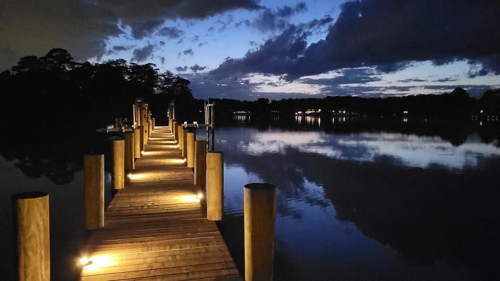 Wooden dock with lights extending into calm water at dusk, reflecting the cloudy sky.
