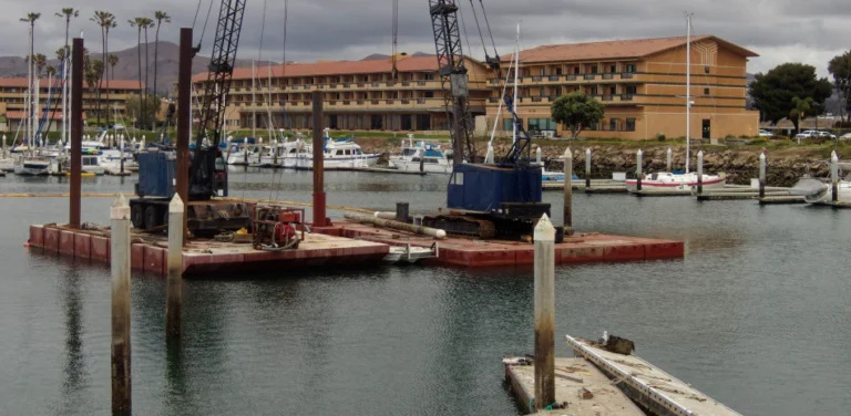 Construction barges in a harbor, with a multi-story building and palm trees in the background.