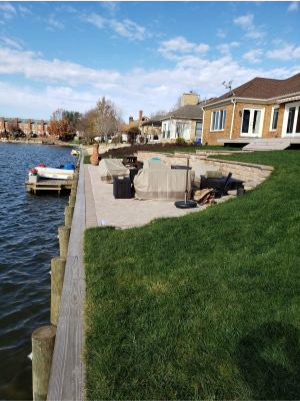 Lakeside backyard with a wooden dock, grassy lawn, and brick patio. Clear sky, sunny day.