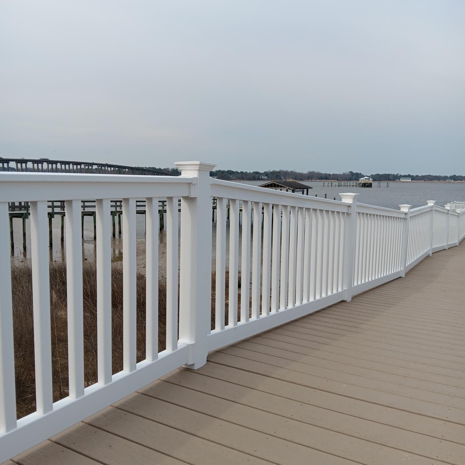 White railing along a boardwalk overlooking water and distant bridges under a cloudy sky.