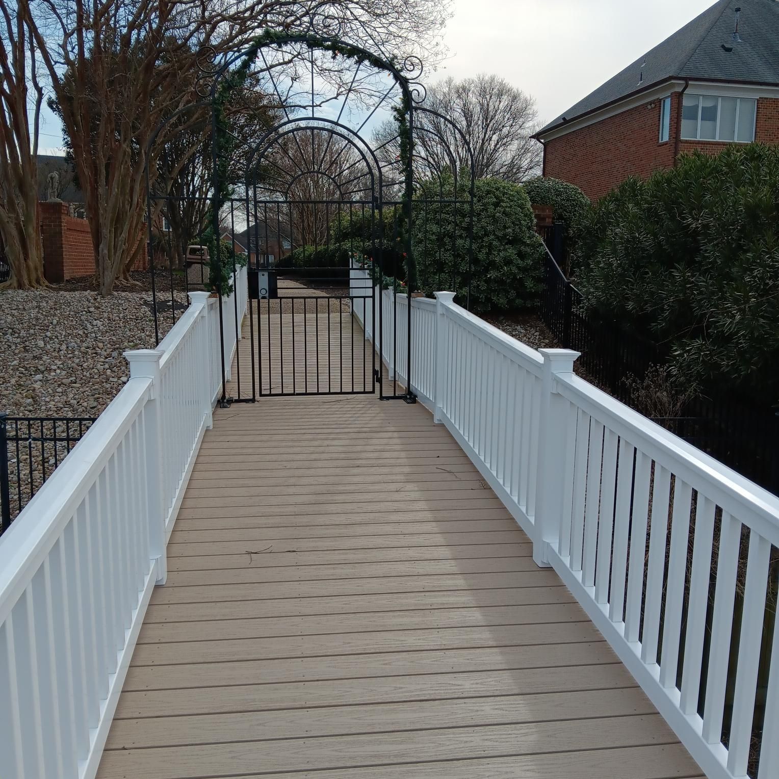 A white-railed bridge with brown decking leads to a black gate under an archway, in a residential area.