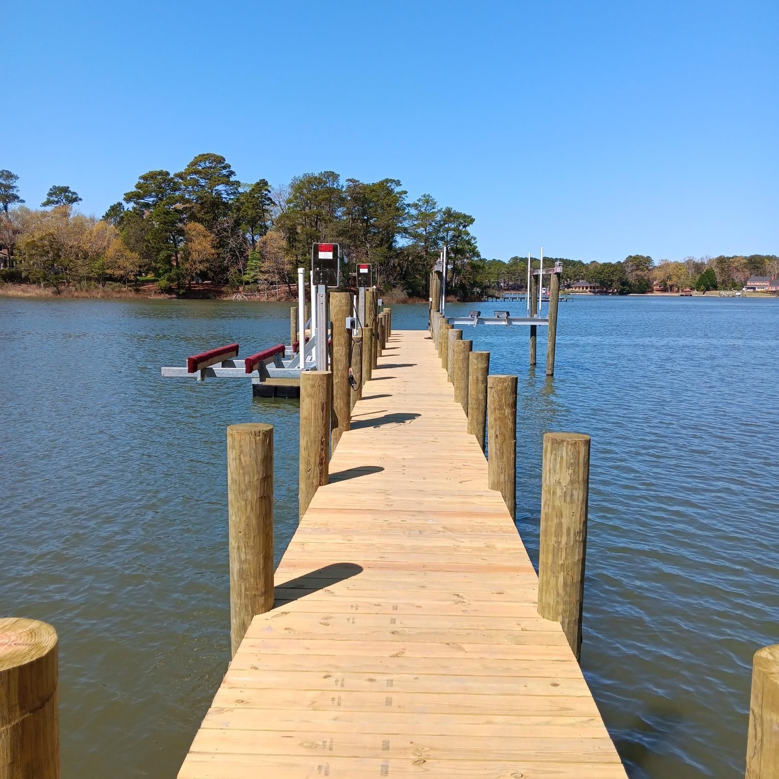 Wooden dock extends over water, leading towards a boat lift and pilings. Blue sky and trees in the background.