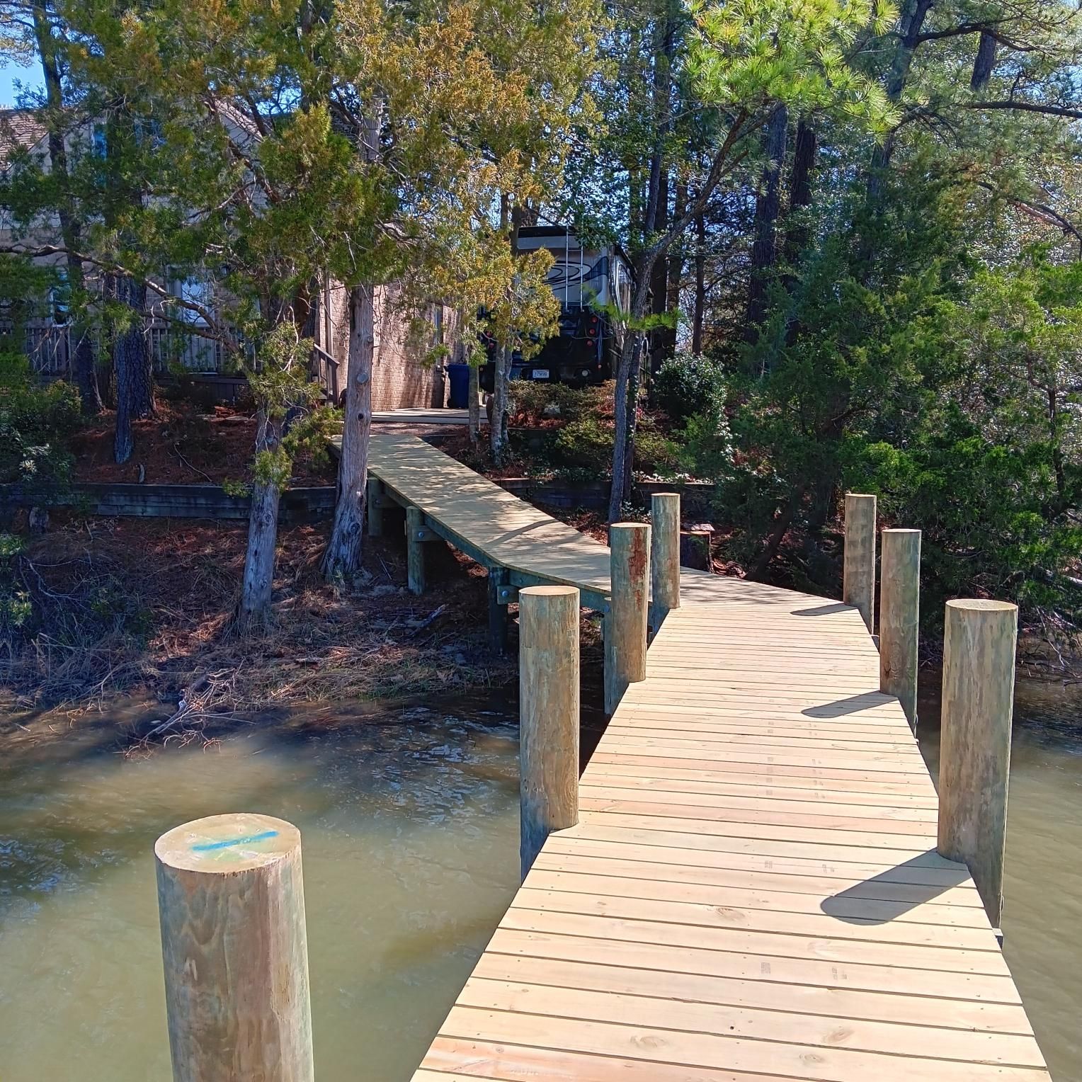 Wooden pier extending over water, leading to a wooded area with trees and a house in the background.