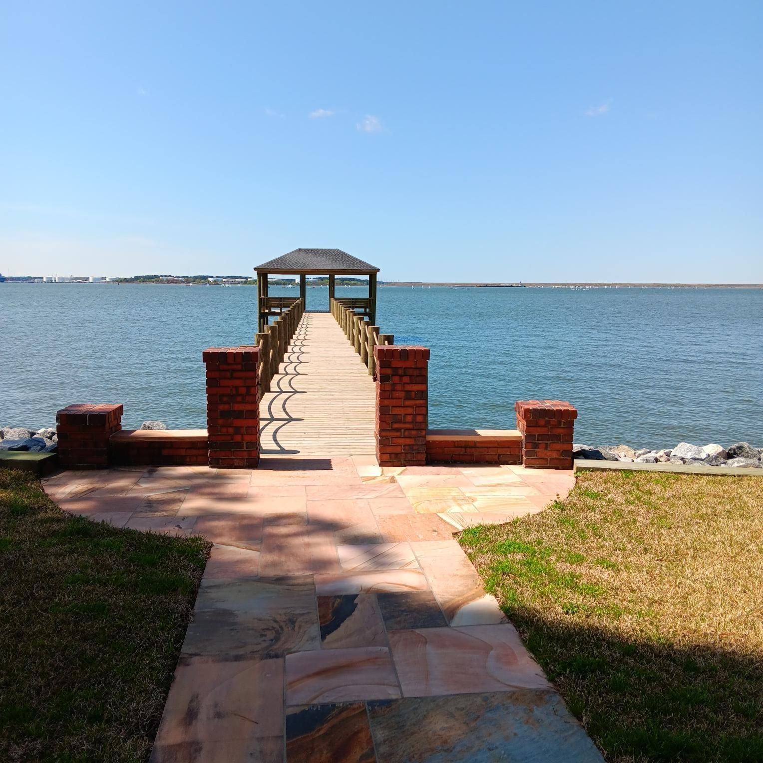 Pathway leading to a wooden pier with gazebo over calm water under a blue sky.