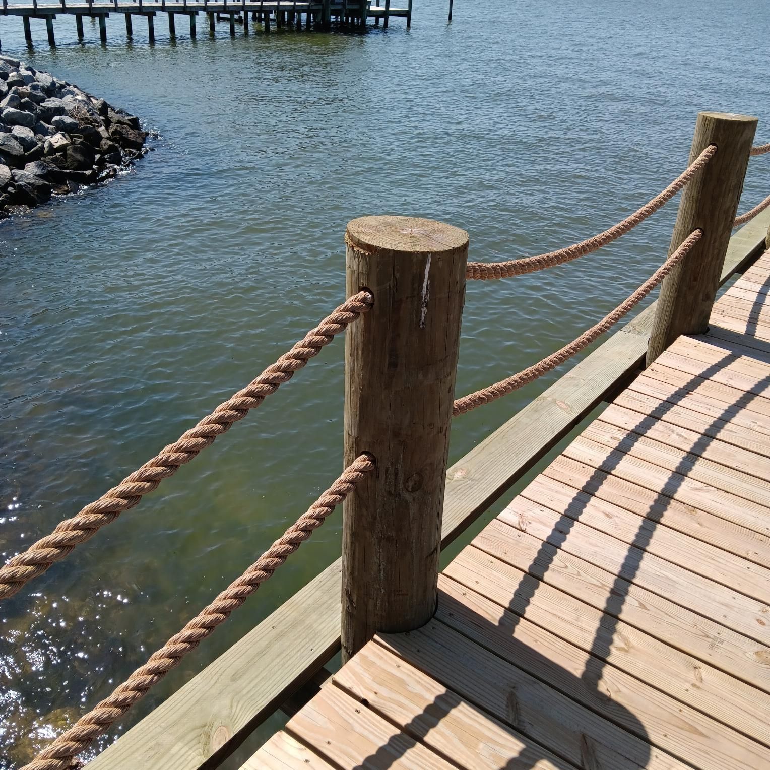 Wooden pier with rope railing overlooking water. Sunlight reflects on the water's surface.