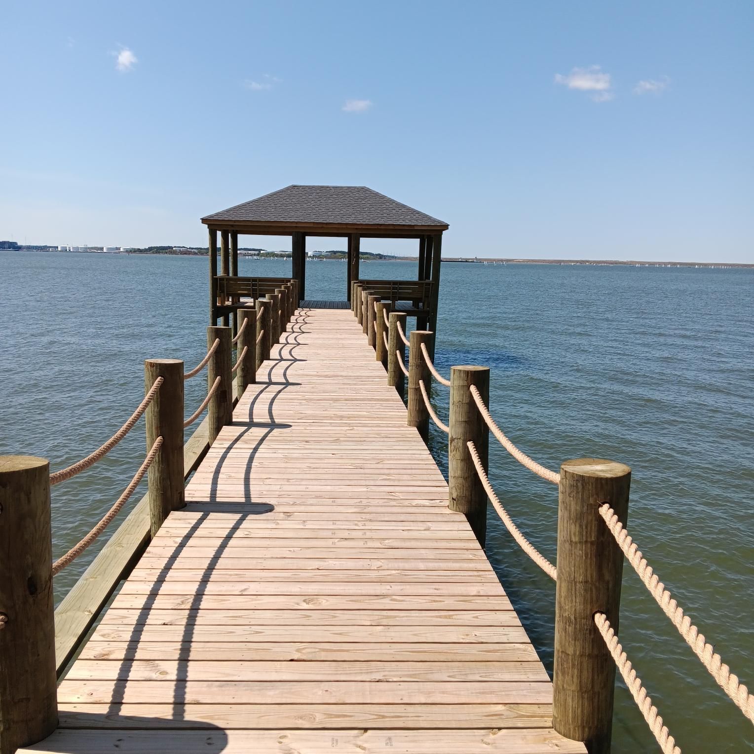 Wooden pier with rope railings and a gazebo at the end, extending into calm blue water under a sunny sky.
