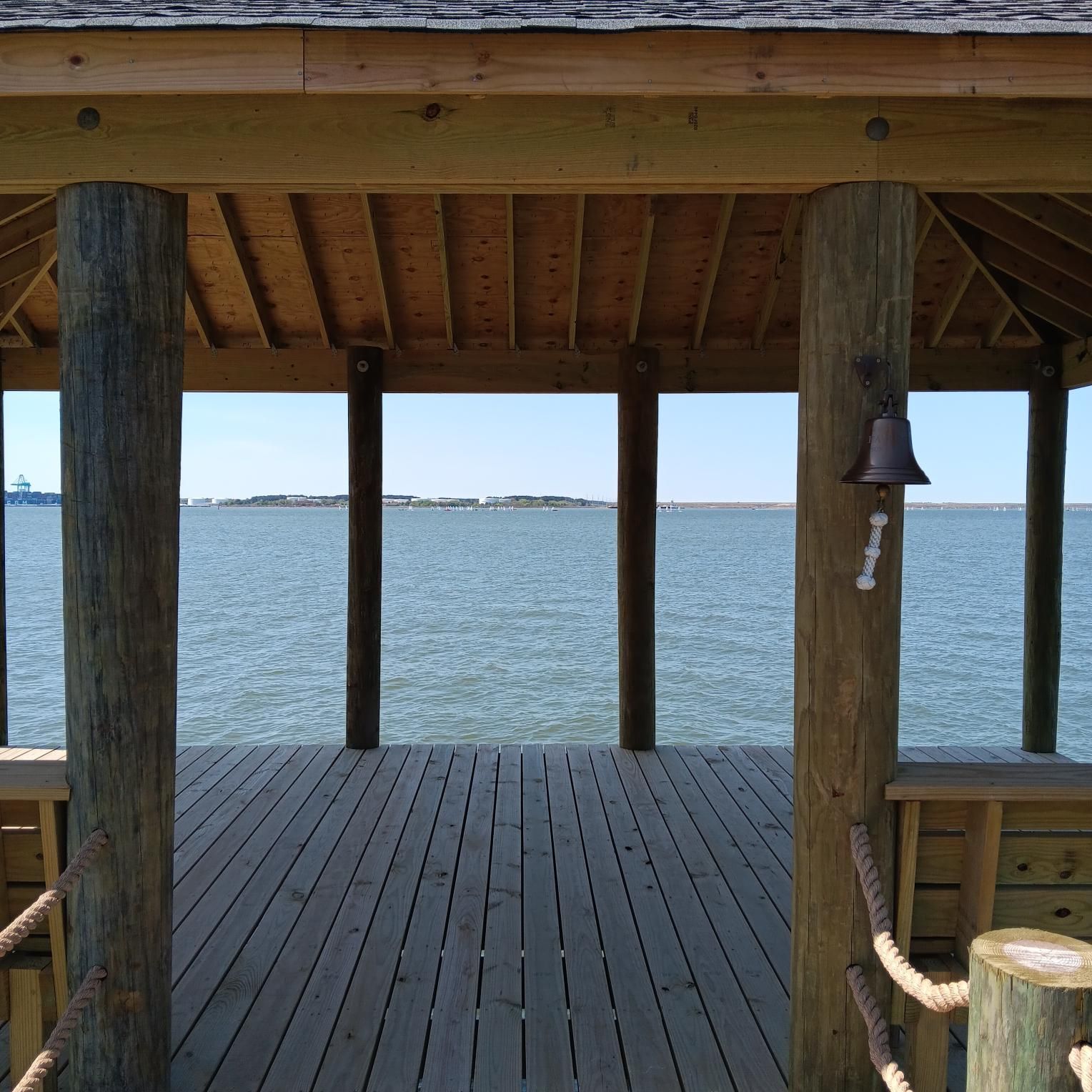 Wooden gazebo on a pier overlooking calm water and distant land. A bell hangs on a support post.