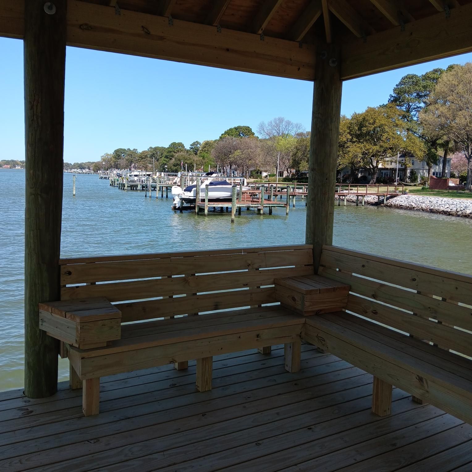 Wooden gazebo with benches overlooking a waterfront with docked boats.