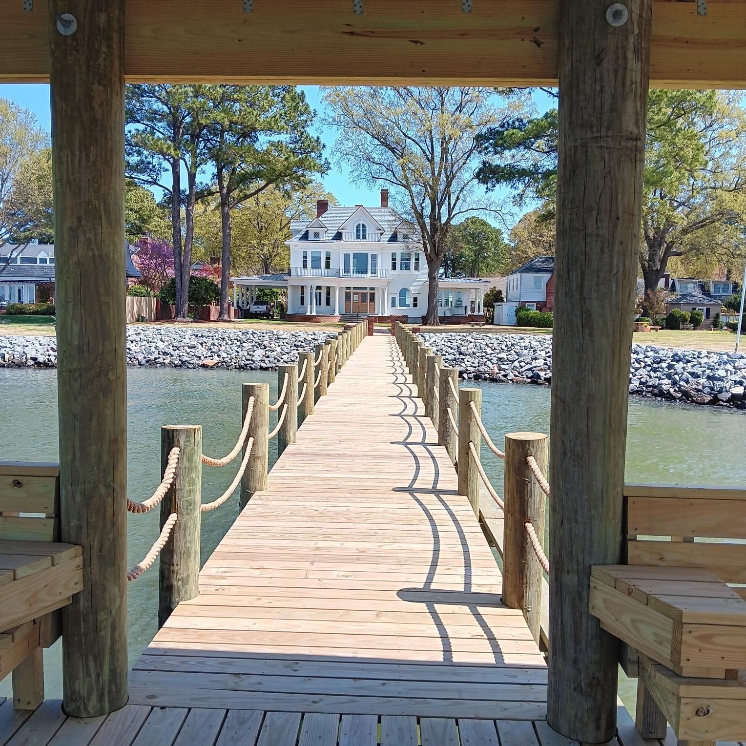 Wooden pier leads to a white house, seen through a shaded structure, beside a body of water on a sunny day.