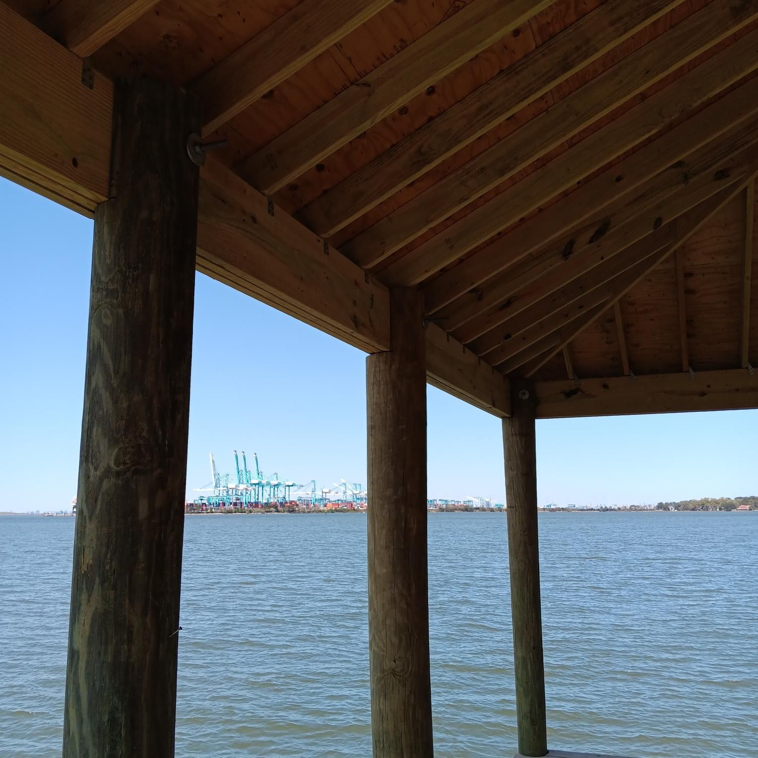 Wooden shelter overlooking water and industrial structures under a blue sky.