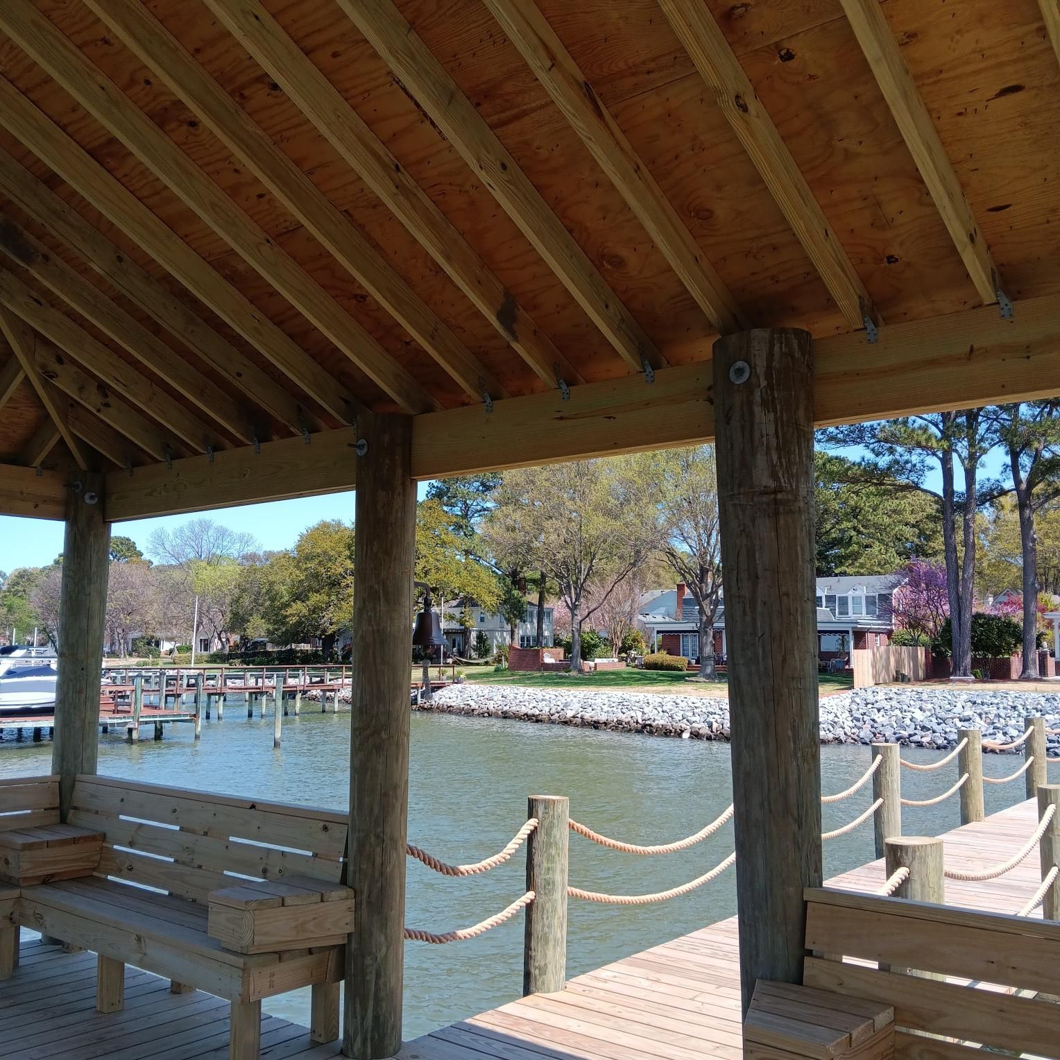 Wooden dock shelter overlooking a calm bay with docked boats and houses in the distance.