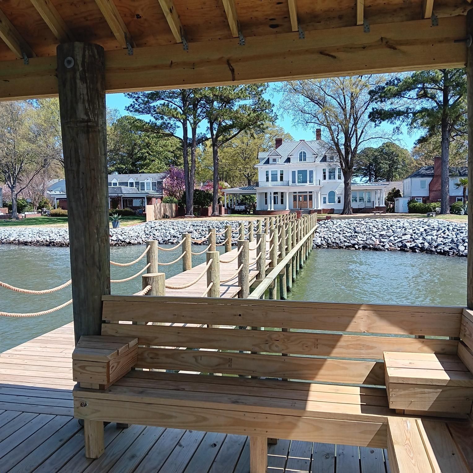 Wooden pier with bench under a covered area, overlooking a large white house and calm water.