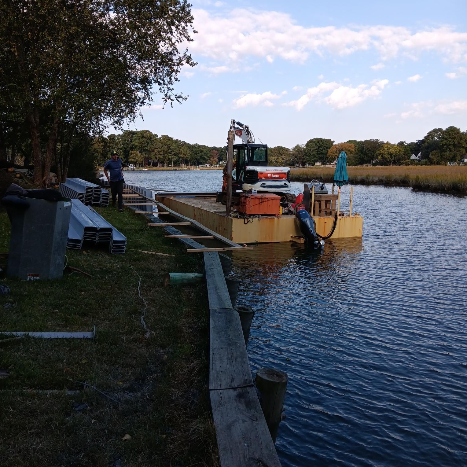 A small excavator on a yellow barge, dredging a waterway. Man stands on the bank, blue sky.