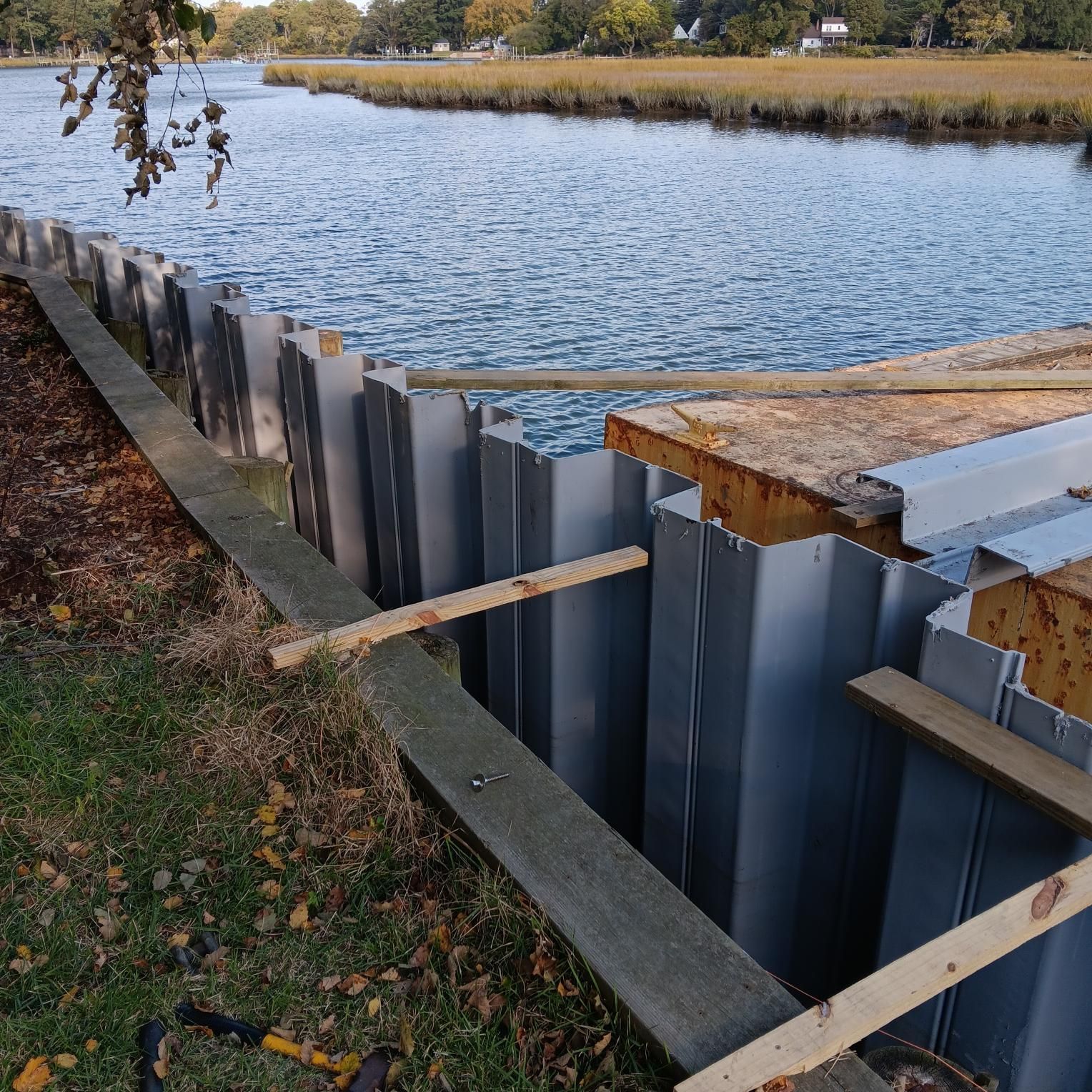 Metal sheet piling installed along a river bank. Boards secured with wooden braces are visible.