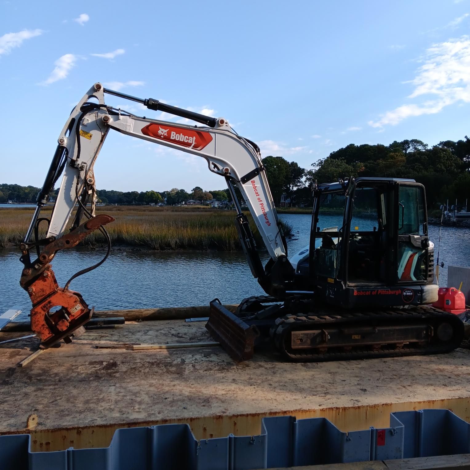 Bobcat excavator on a platform near water, using a hydraulic breaker attachment.